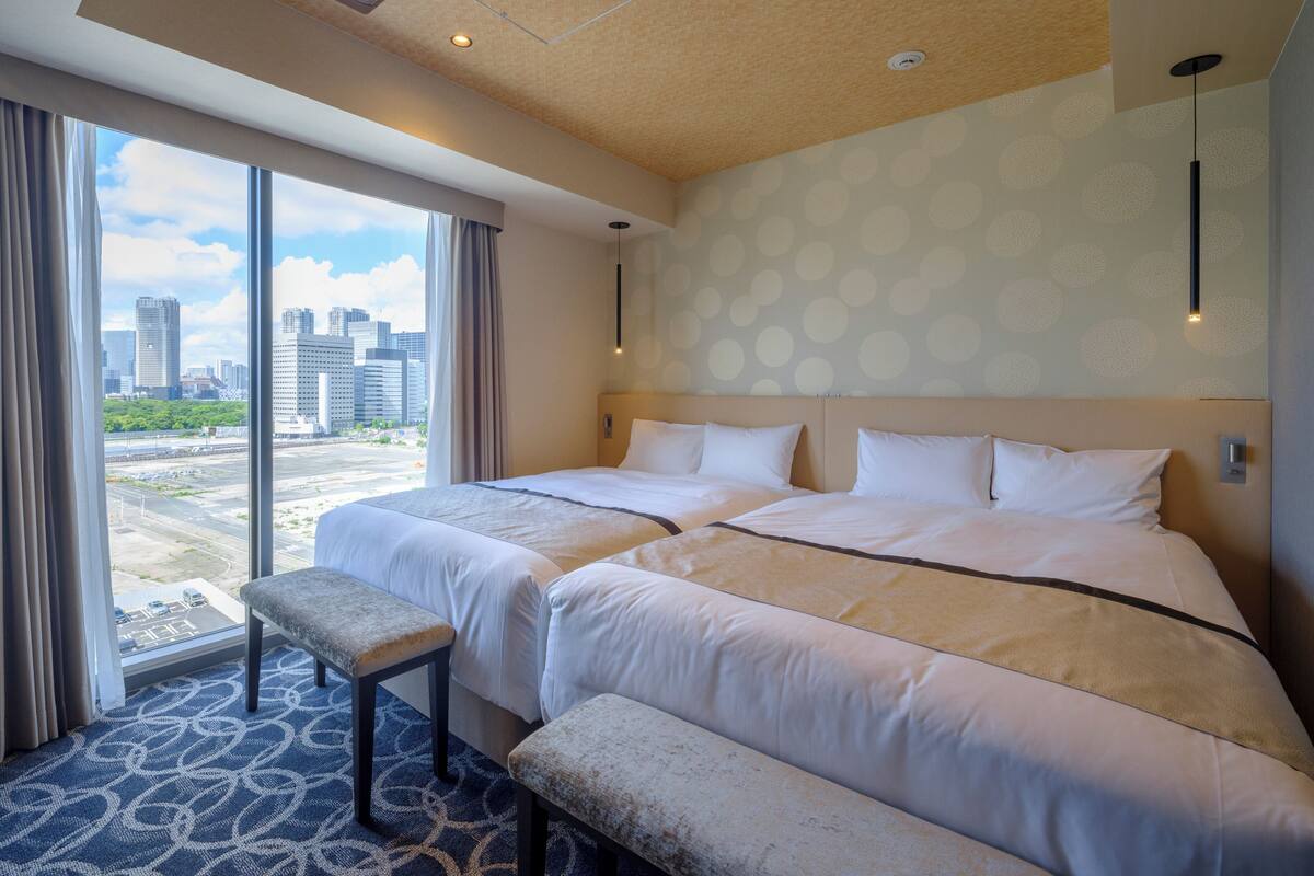 A hotel room with two beds, a bench, and a large window offering a city view.