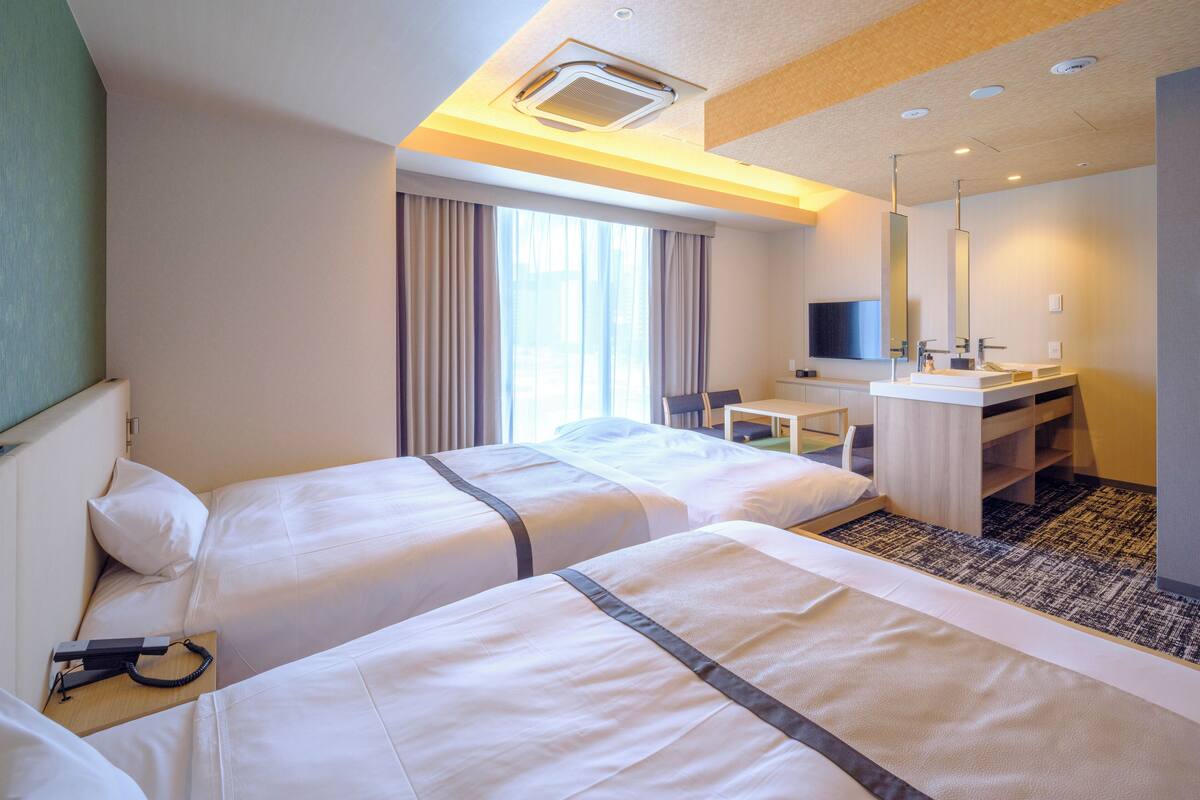 A hotel room with two beds, a desk, a chair, a TV, and a bathroom with a mirror and sink.