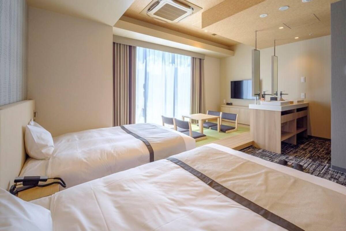 A hotel room with two beds, a desk, a chair, a TV, and a bathroom with a mirror and sink.