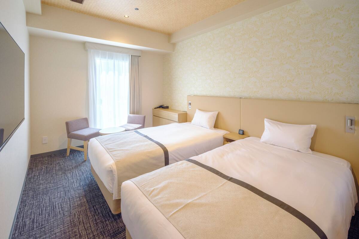 A hotel room with two beds, a desk, a chair, and a large window with curtains.