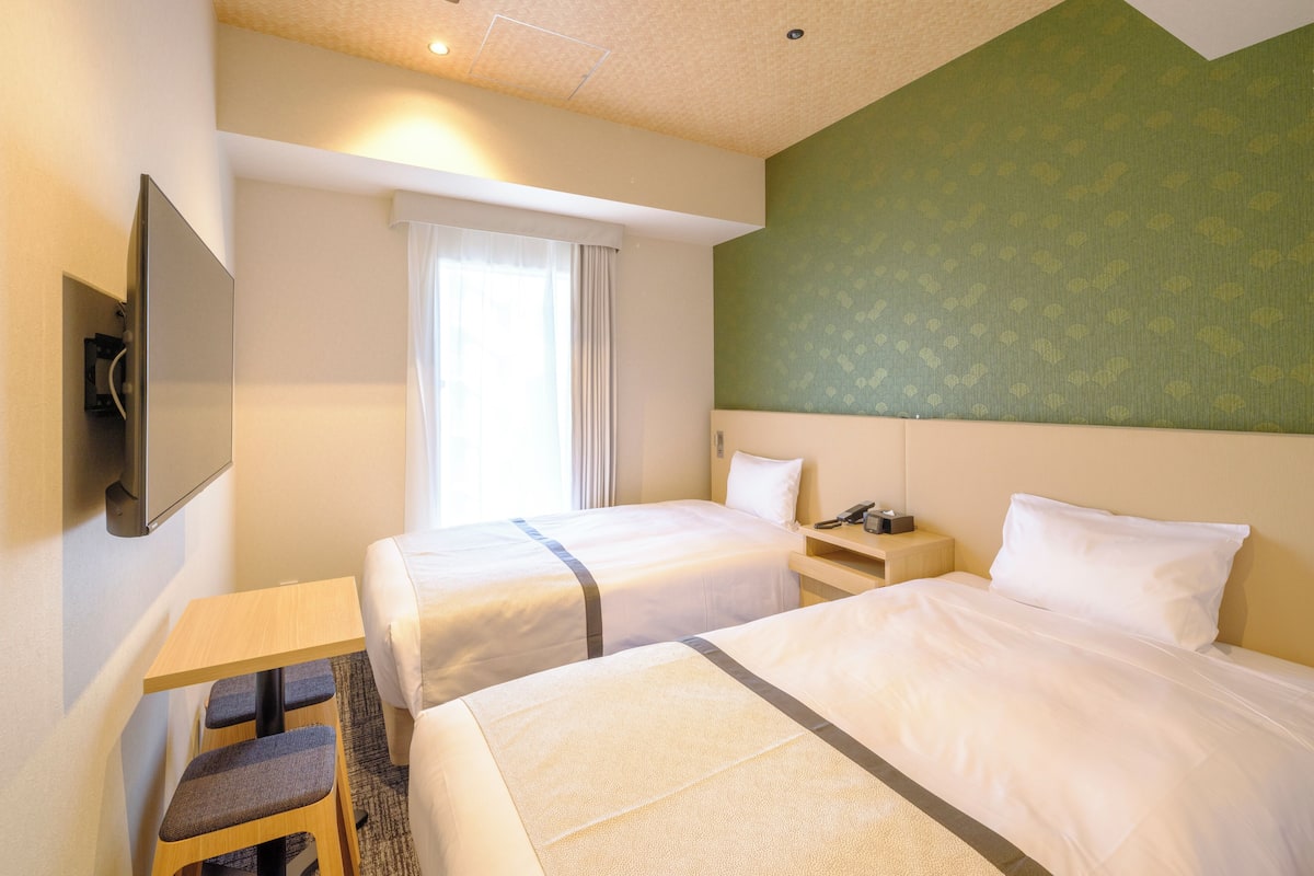 A hotel room with two beds, a small table, a chair, a TV, and a window with curtains.