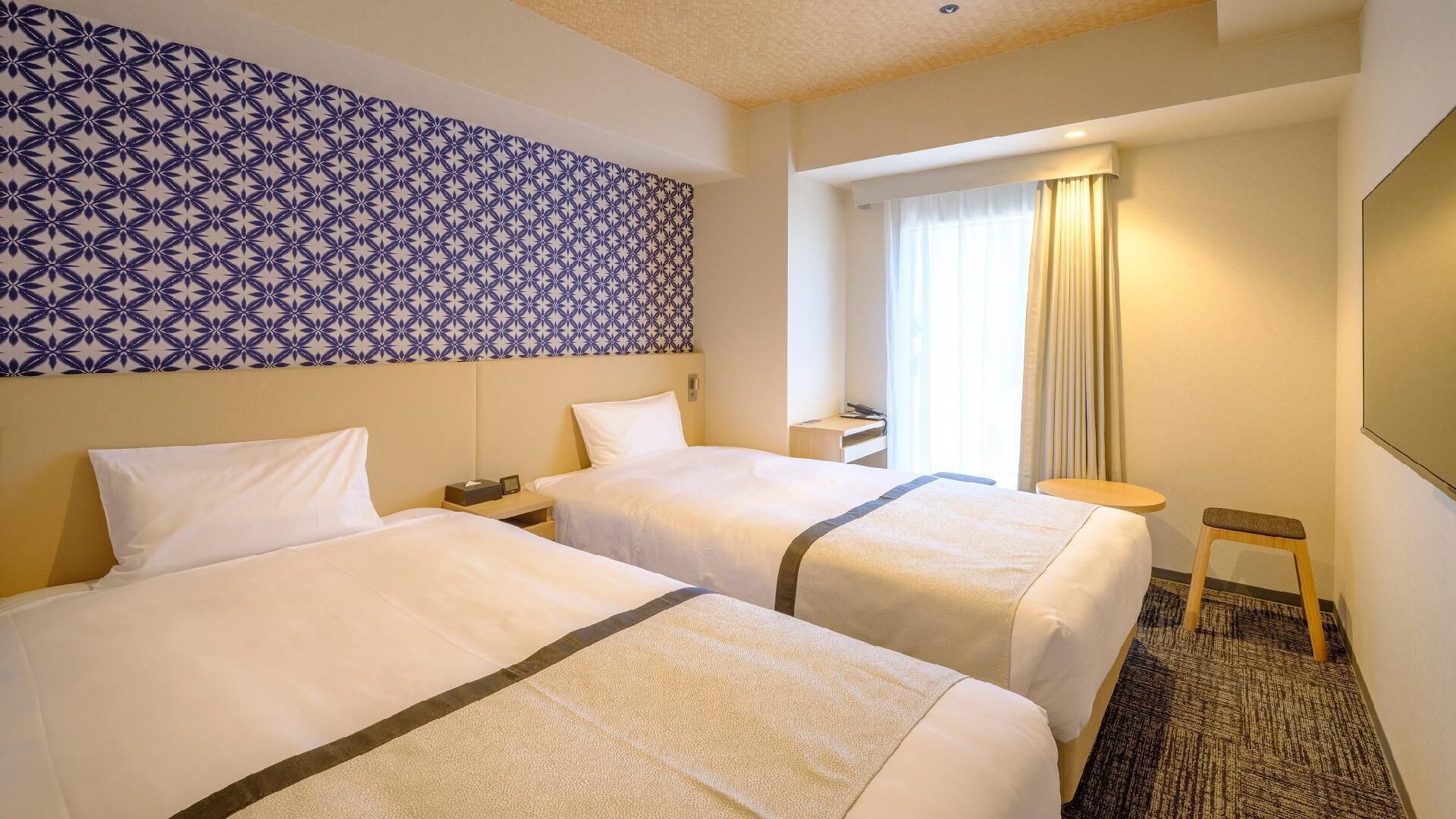 A hotel room with two beds, a patterned wall, a window with curtains, and a small table.