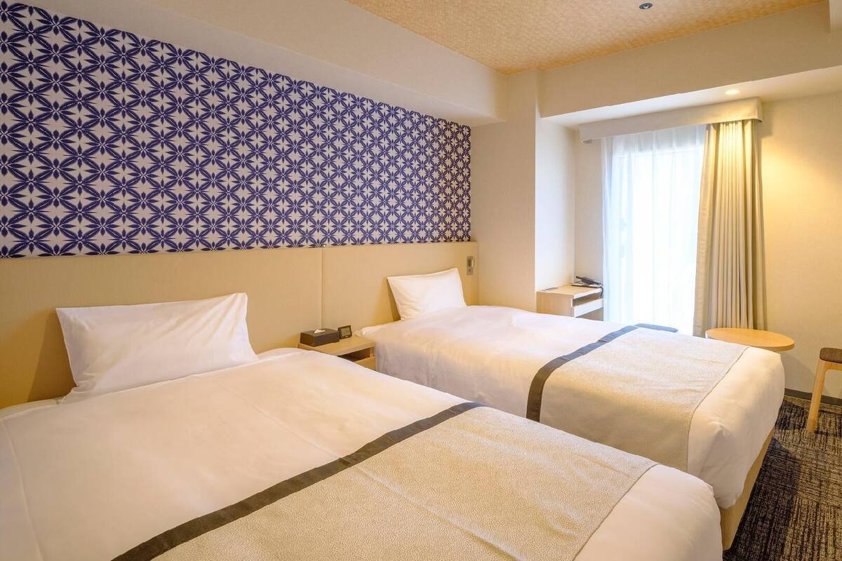 A hotel room with two beds, a patterned wall, a window with curtains, and a small table.