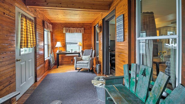 A cozy cabin interior with wooden walls, a fireplace, a rocking chair, and a large window with curtains. - Northville Lakefront Escape w/ Deck: Walk to Lake (Northville)