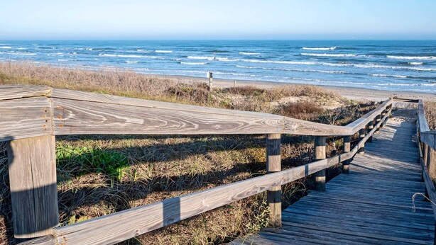 Ein hölzerner Steg führt zum Strand, im Hintergrund schlagen die Wellen des Ozeans.