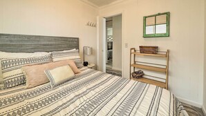 A bedroom with a bed, pillows, a nightstand, a lamp, a wooden shelf, and a green-framed window. - Lakefront Cottage Near Wineries + State Parks! (Mears)
