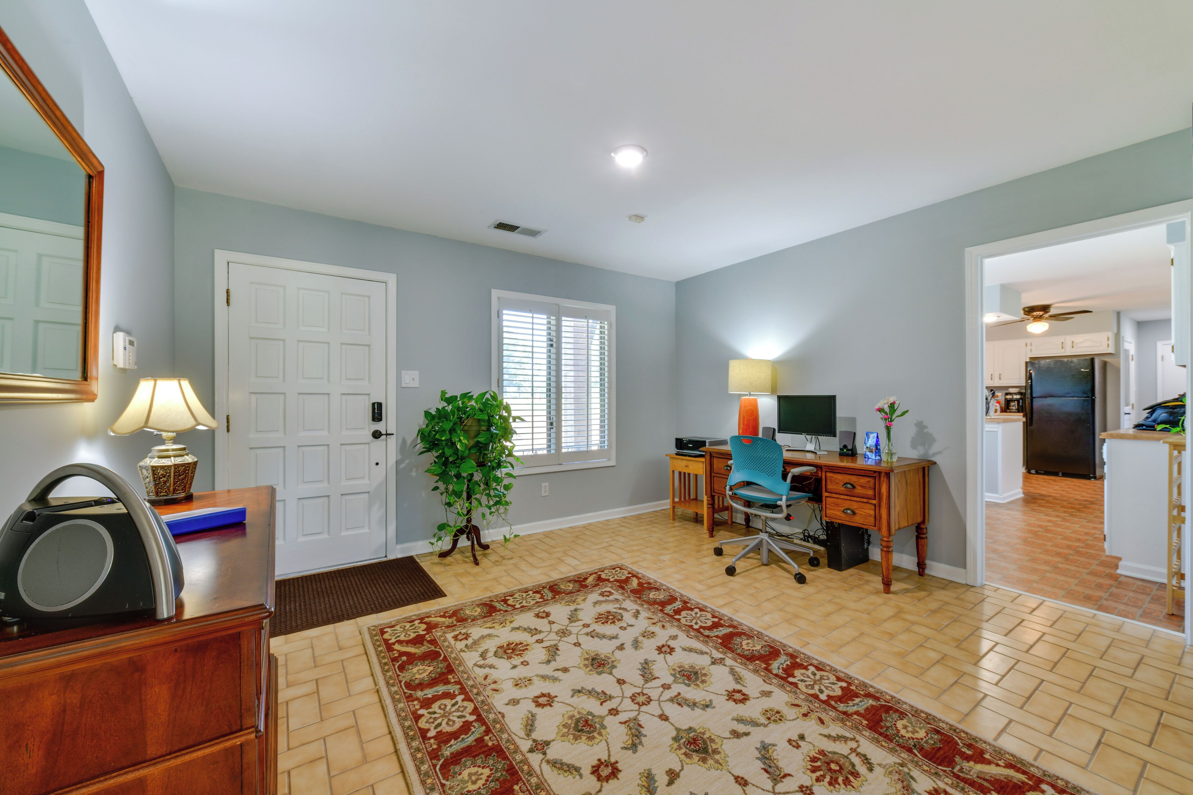 A spacious living room with a large rug, a desk with a computer, a plant, and a door leading to another room.
