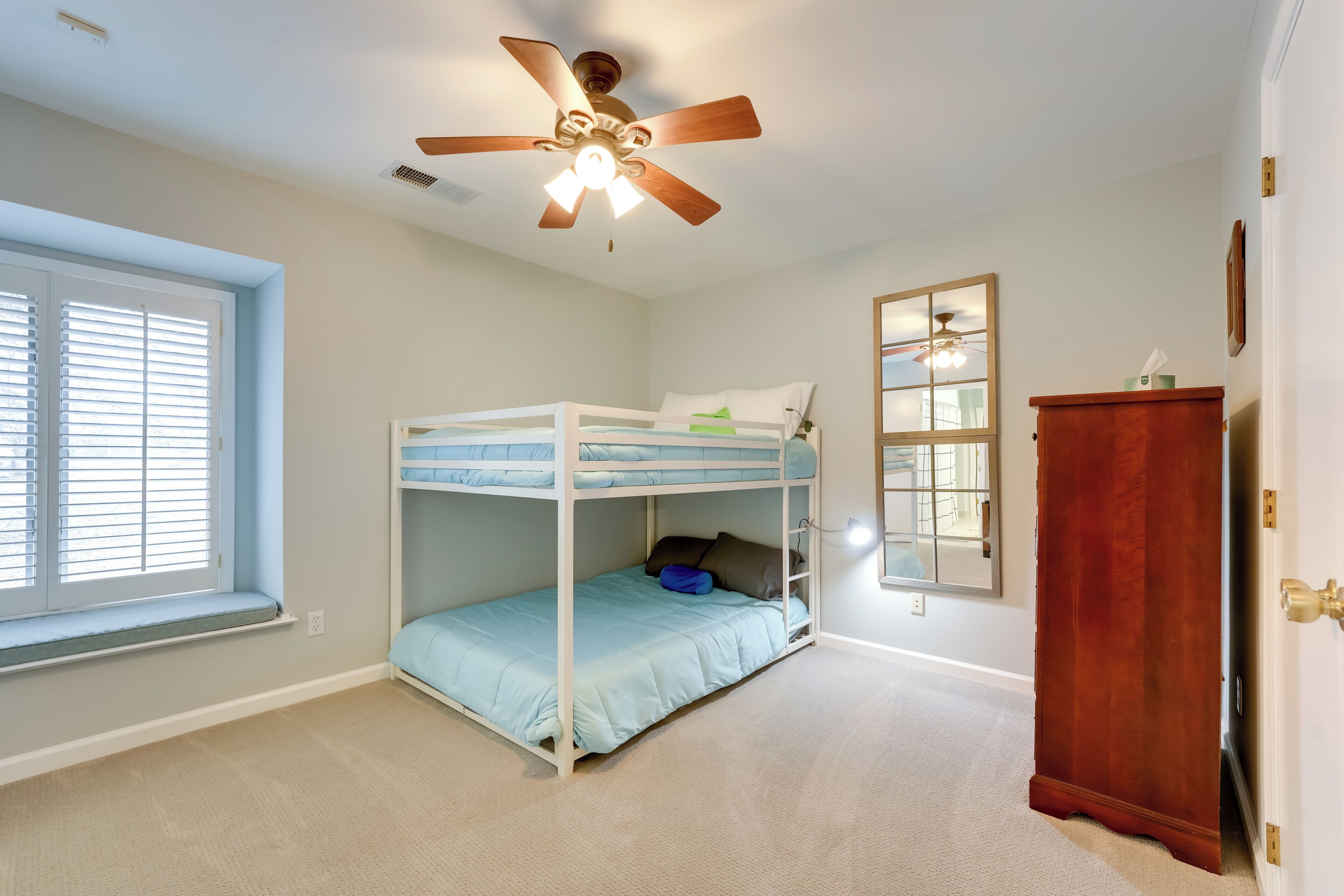 Une chambre avec un lit superposé, un ventilateur de plafond, une fenêtre avec des stores, une armoire en bois et un miroir au mur.