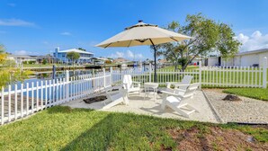 A waterfront patio with white chairs and a table under a large umbrella, surrounded by a white picket fence and a clear view of the water. - Canal-front Haven: Pool, Dock & Lanai in Palmetto (Palmetto)