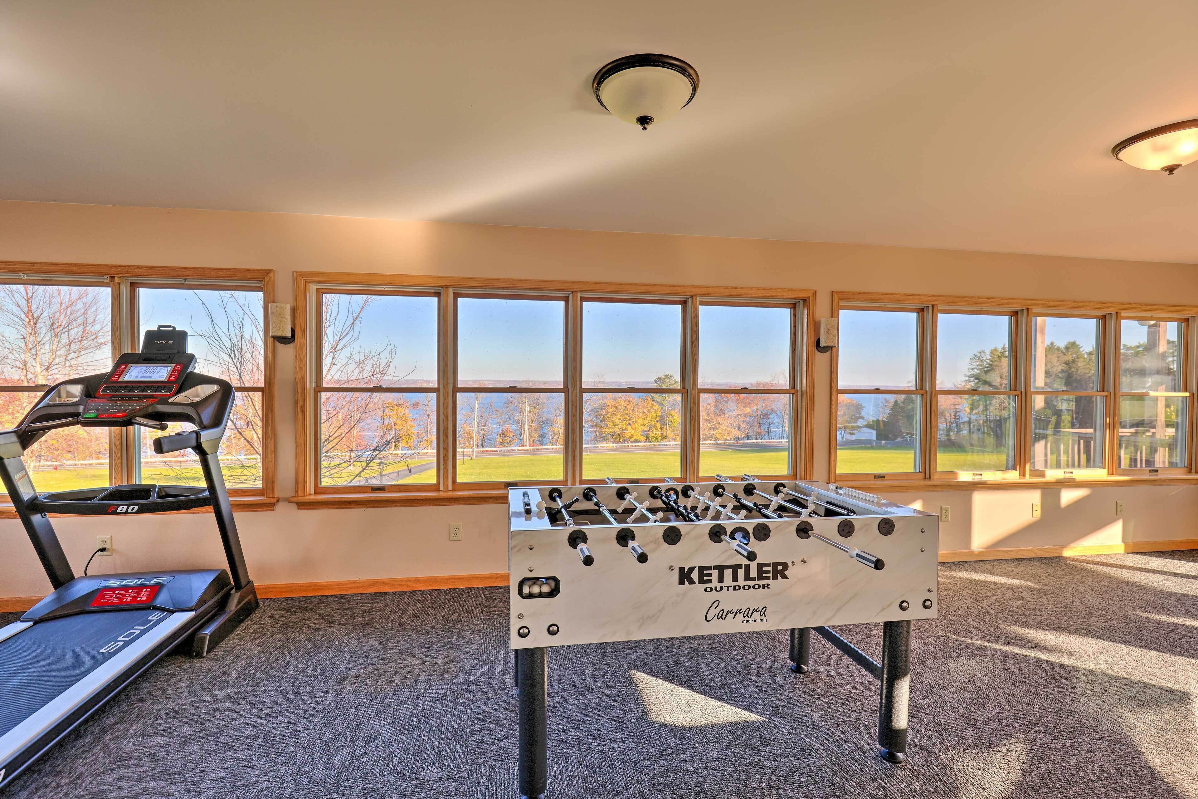 A room with a foosball table, a treadmill, and large windows with a view of a park.