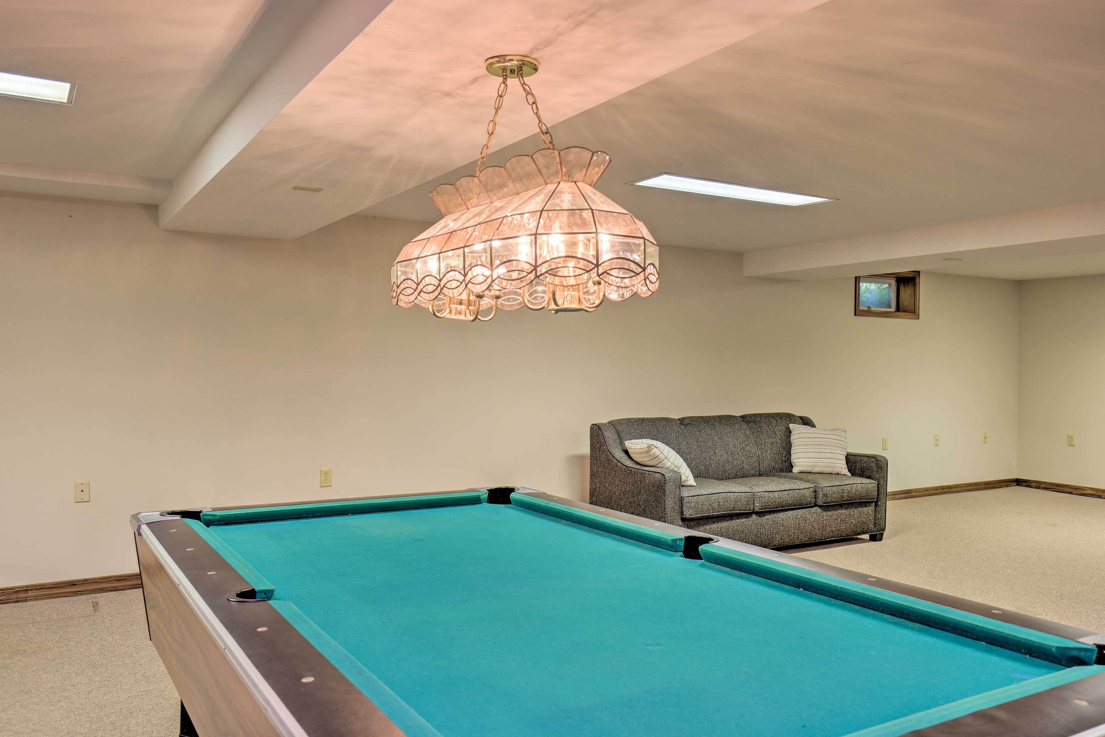 Une table de billard dans une pièce dotée d’un ventilateur de plafond, d’un petit téléviseur et d’un canapé gris.