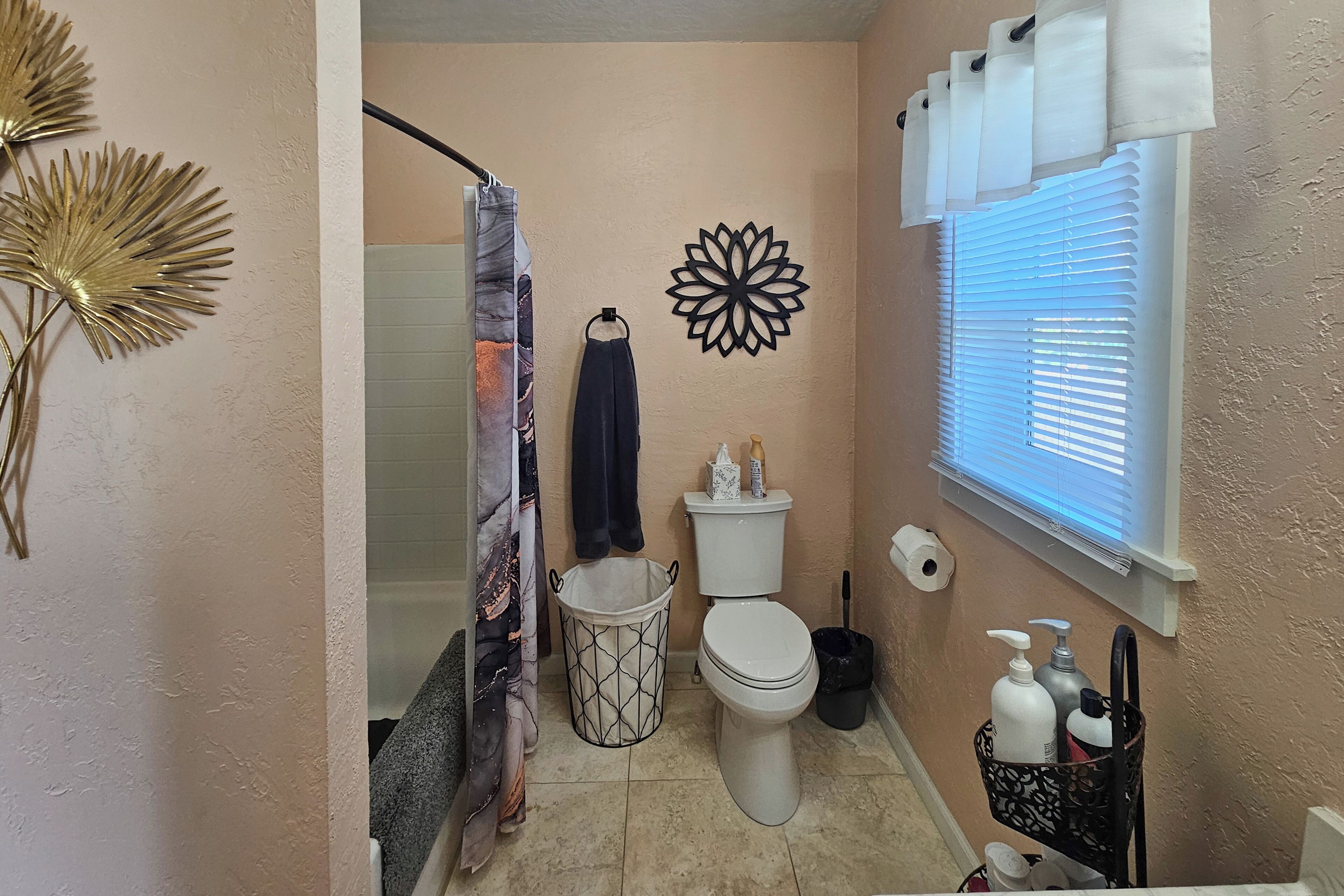A bathroom with a toilet, a shower curtain, a wall-mounted towel rack, a window with blinds, and a trash bin.