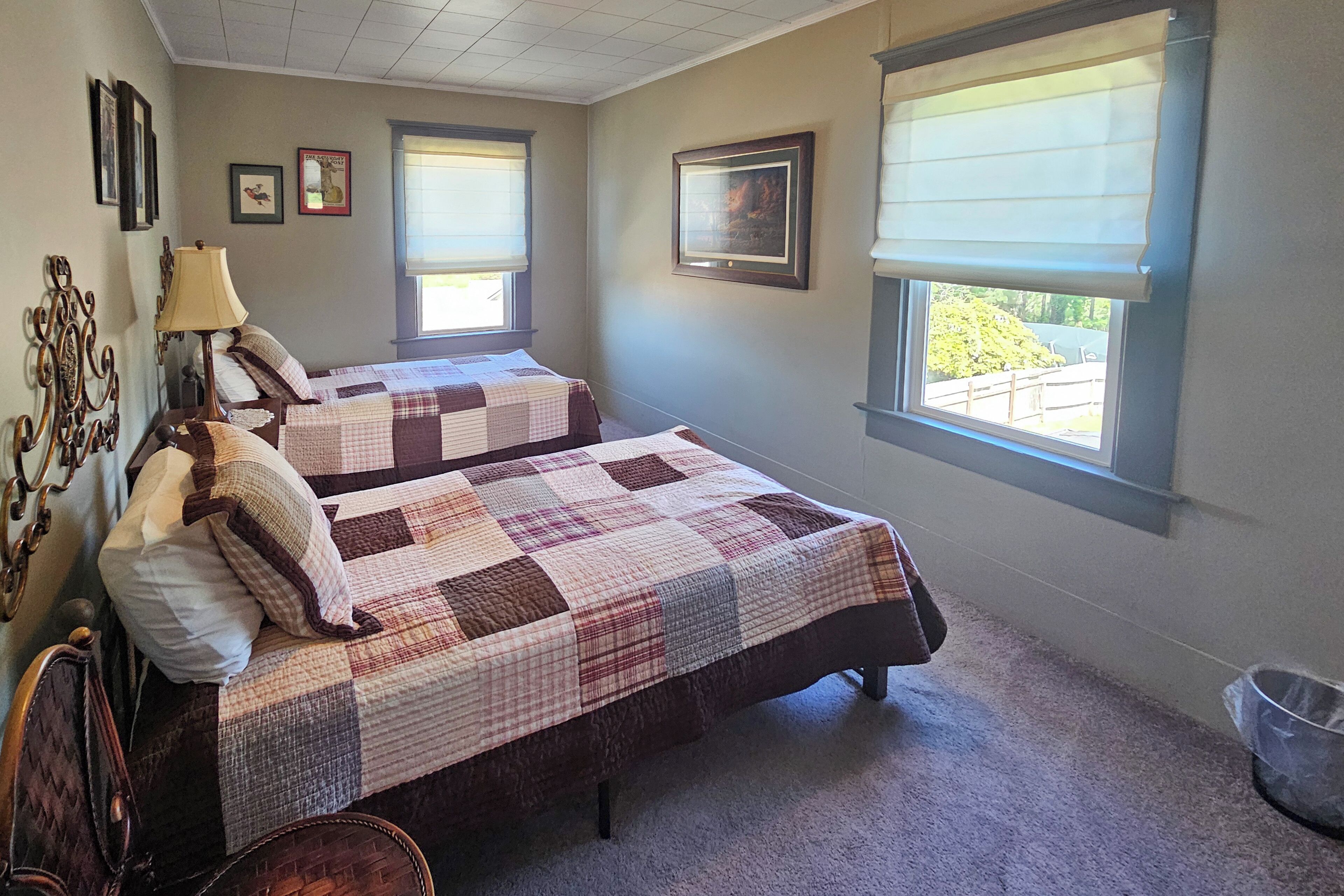 A bedroom with two beds, a window with blinds, a lamp, and a framed picture on the wall.