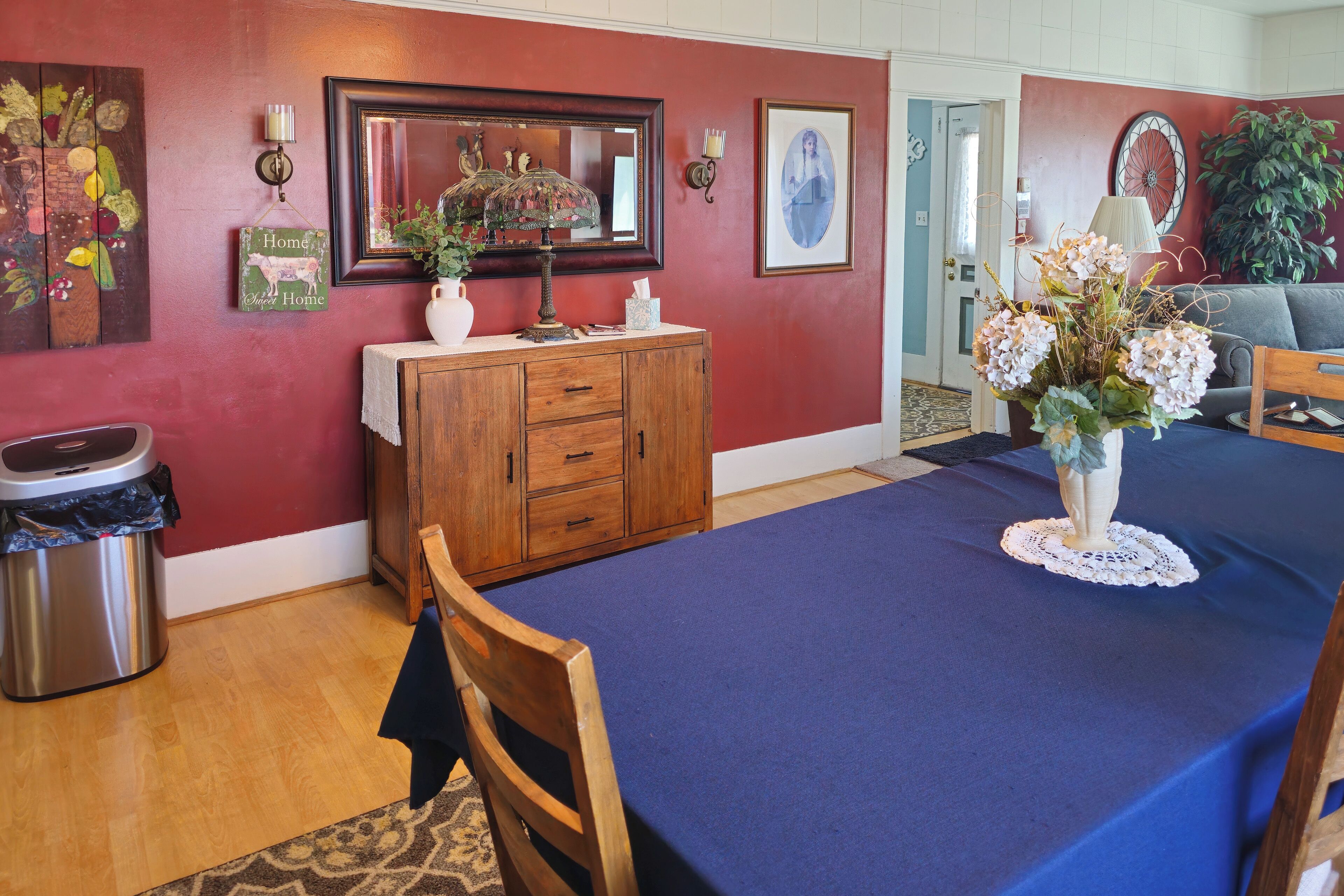 A dining area with a blue tablecloth, wooden chairs, a vase of flowers, a wooden cabinet, and wall decorations.