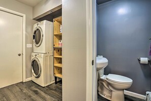 A laundry room with stacked washers and dryers, a wooden shelf unit, and a toilet in a bathroom with dark walls.