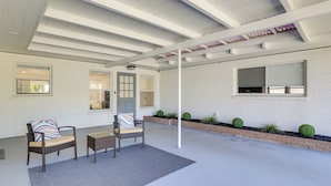 A covered patio area with two wicker chairs and a small table, a white wall with windows, and a brick planter with greenery. - Dog-friendly Home w/ Patio & BBQ in Bradenton (Bradenton)