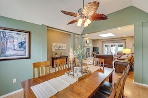 A dining area with a wooden table, chairs, and a ceiling fan. There is a fireplace in the background and a painting on the wall.