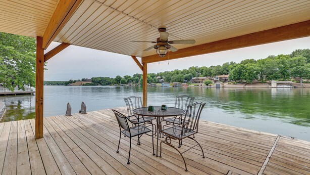 A wooden deck with a round table and chairs, a ceiling fan, and a view of a lake with houses in the background. - Bella Vista Home w/ Kayaks & Deck on Loch Lomond! (Bella Vista)