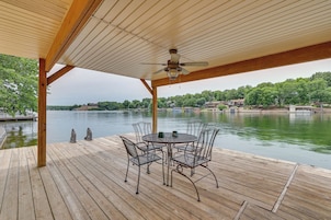 A wooden deck with a round table and chairs, a ceiling fan, and a view of a lake with houses in the background.