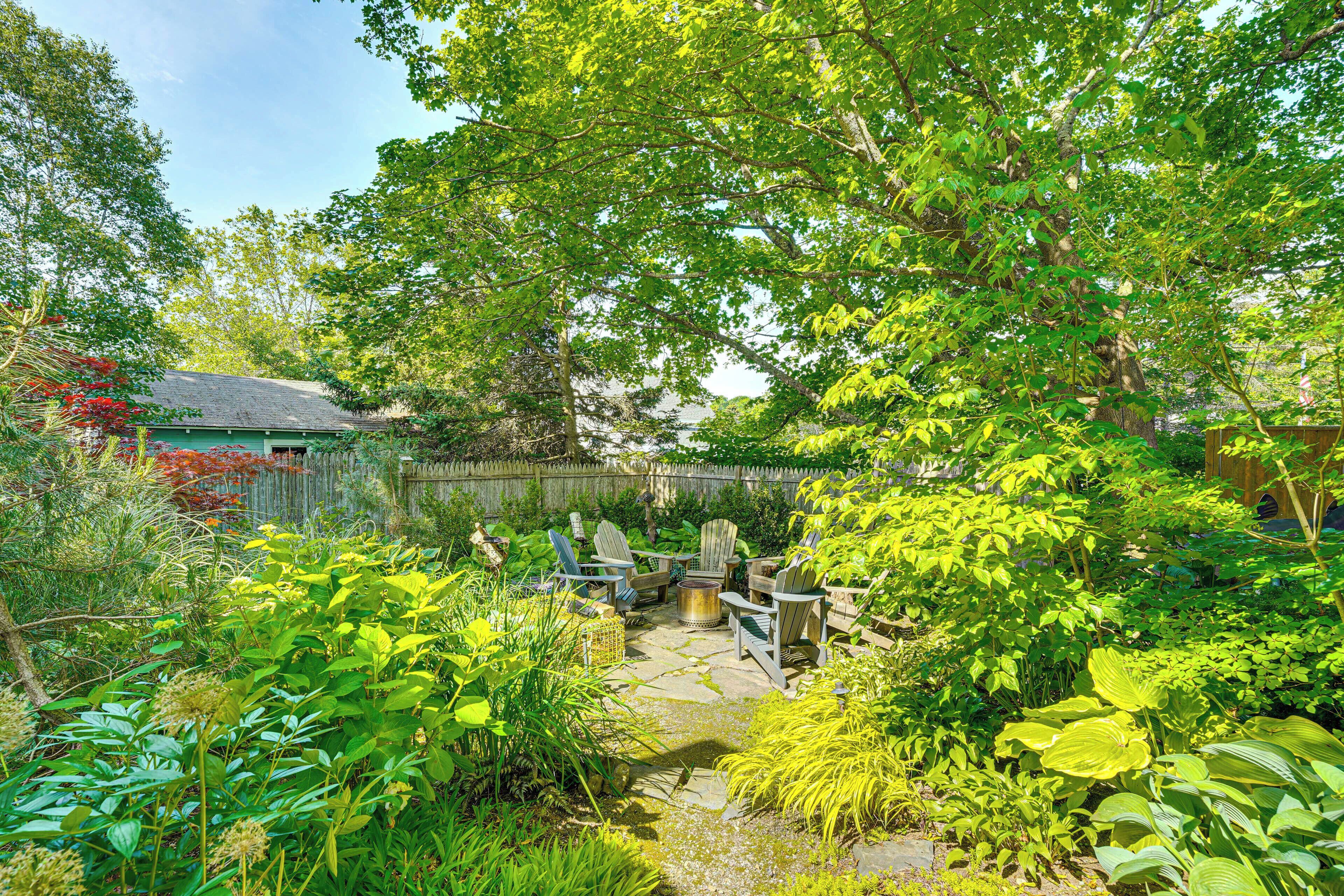 A garden with a stone patio, wooden chairs, and lush greenery.