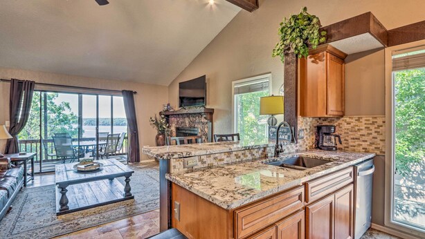 A spacious living area with a large window, a fireplace, and a kitchen with a granite countertop and wooden cabinets. - 1 Mi to Margaritaville! Cabin w/ Resort Perks (Osage Beach)