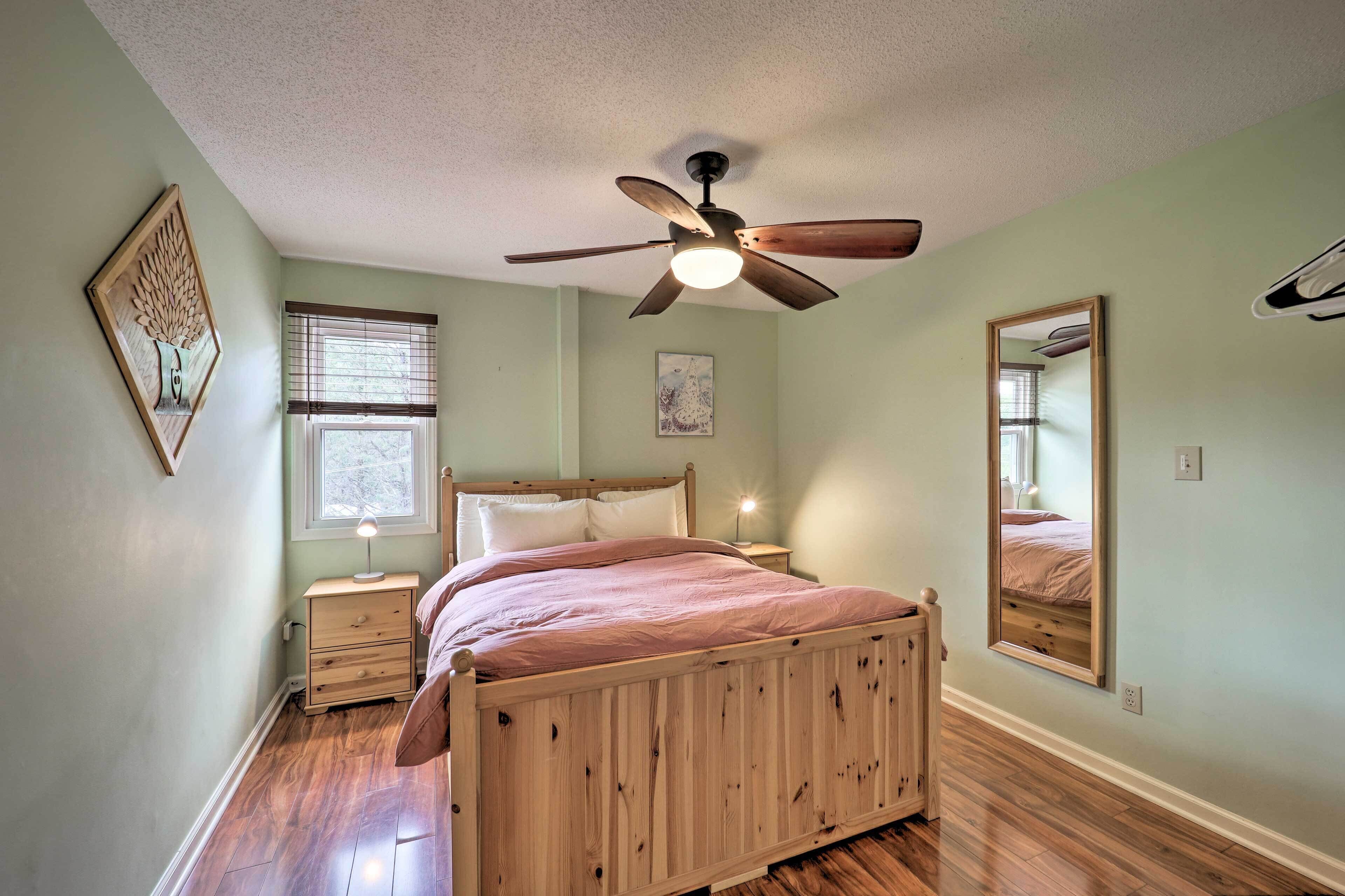 A bedroom with a wooden bed, a ceiling fan, a nightstand, a bedside lamp, a window with blinds, and a mirror on the wall.