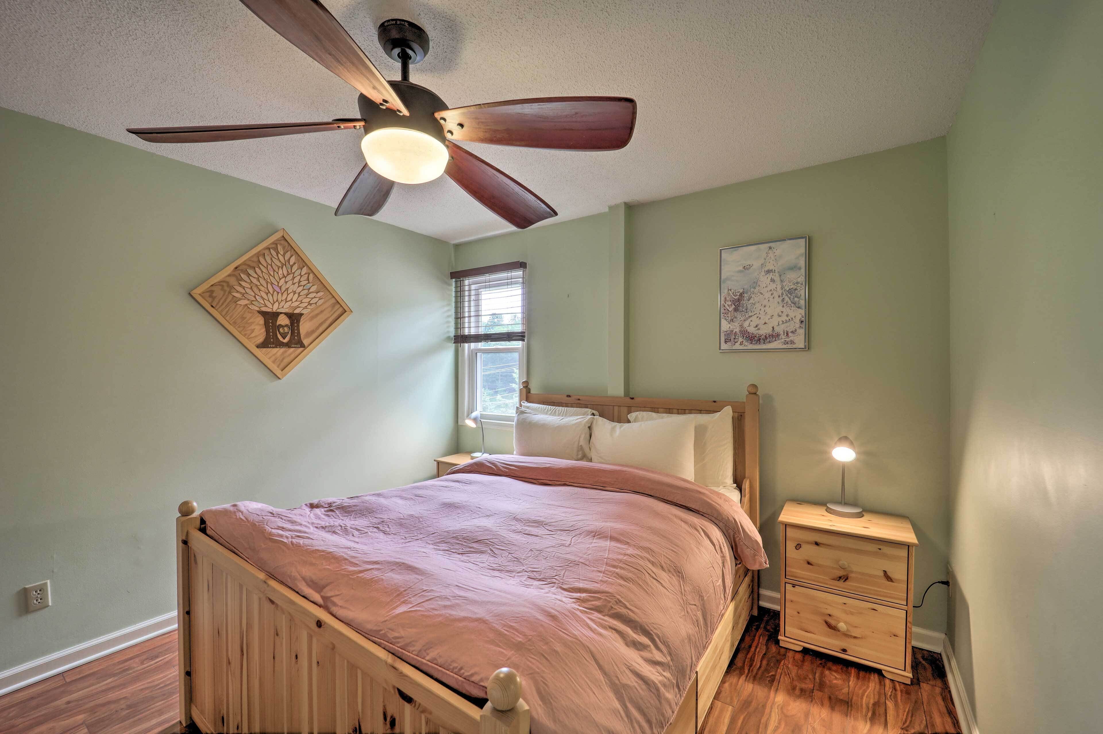 A bedroom with a wooden bed, a ceiling fan, a nightstand with a lamp, and artwork on the walls.