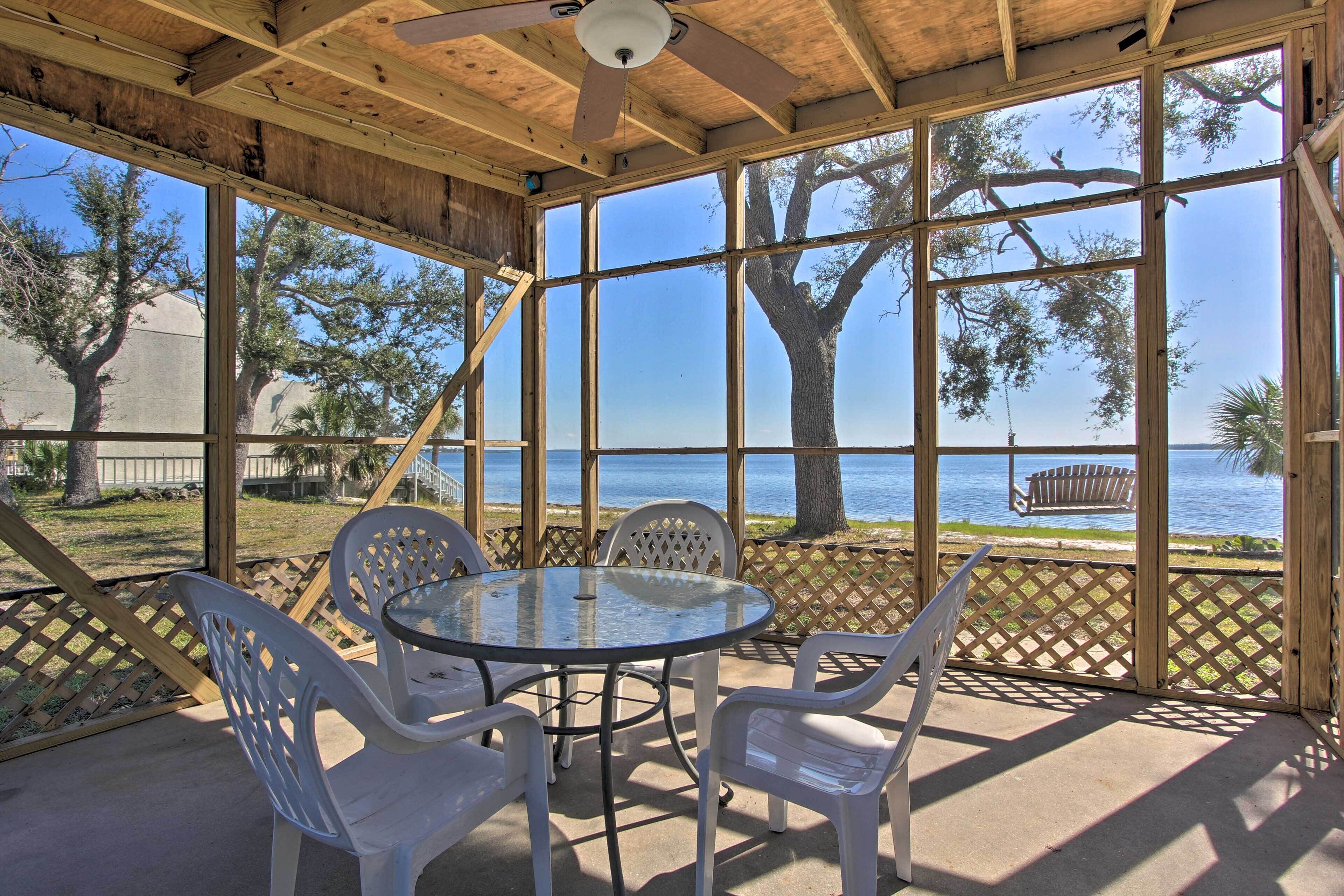 A covered patio with a round glass table and white plastic chairs, overlooking a body of water and trees.