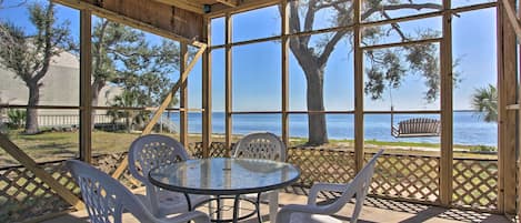 Un patio techado con una mesa redonda de vidrio y sillas de plástico blancas, con vistas a un cuerpo de agua y árboles.
