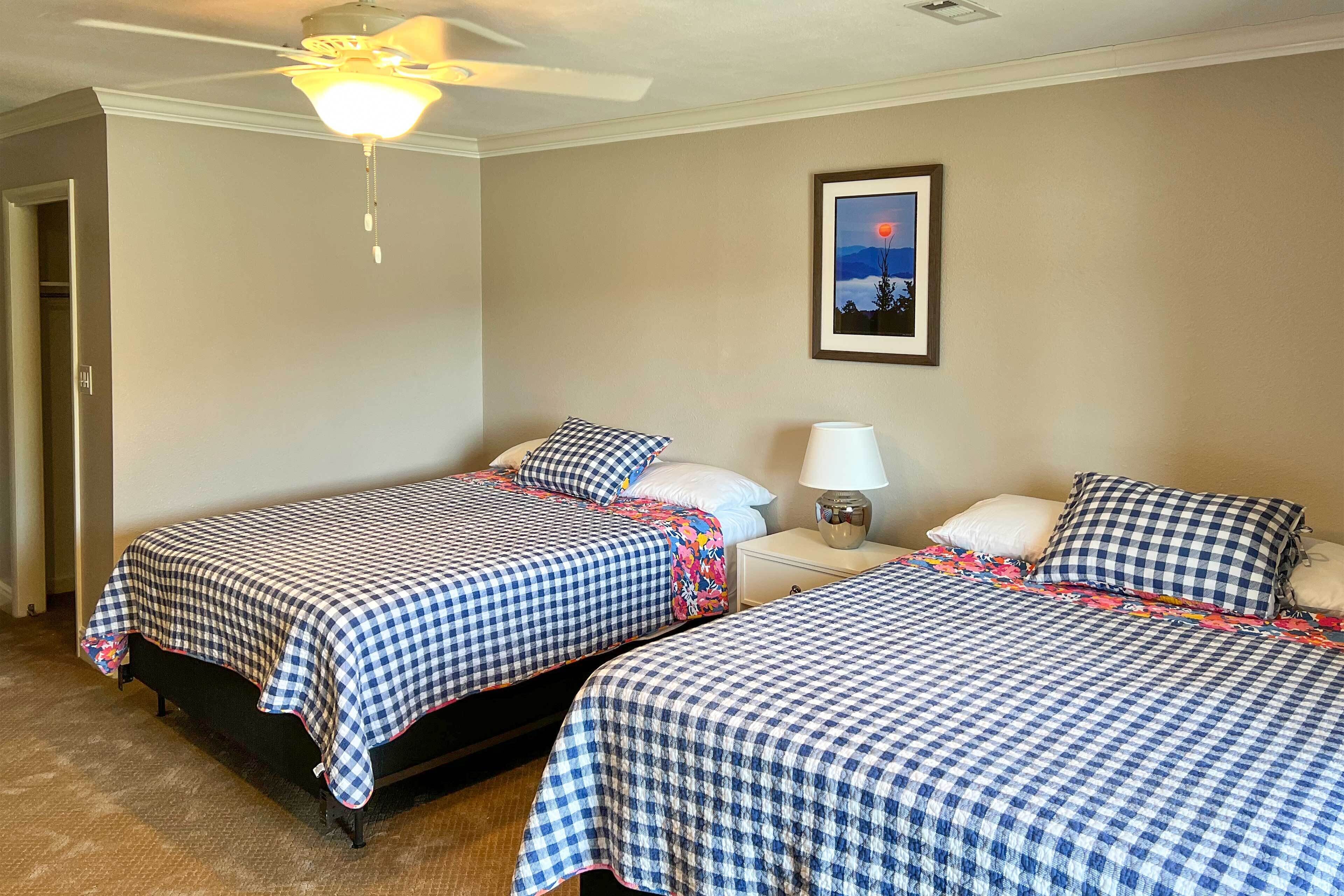 Habitación de hotel con dos camas, un ventilador de techo, una mesita de noche con lámpara y un cuadro en la pared.