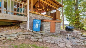 A rustic outdoor area with a stone wall, a blue barrel labeled "PUTCANS HERE," and a grill. - Troutfest: Anglers Retreat on White River w/ Deck (Cotter)