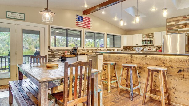 A kitchen with a wooden table, bar stools, and a large window overlooking greenery. - Troutfest: Anglers Retreat on White River w/ Deck (Cotter)