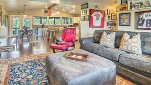 A spacious living room with a gray sofa, a red chair, and a coffee table. There are framed sports memorabilia on the walls and a kitchen in the background. - Troutfest: Anglers Retreat on White River w/ Deck (Cotter)