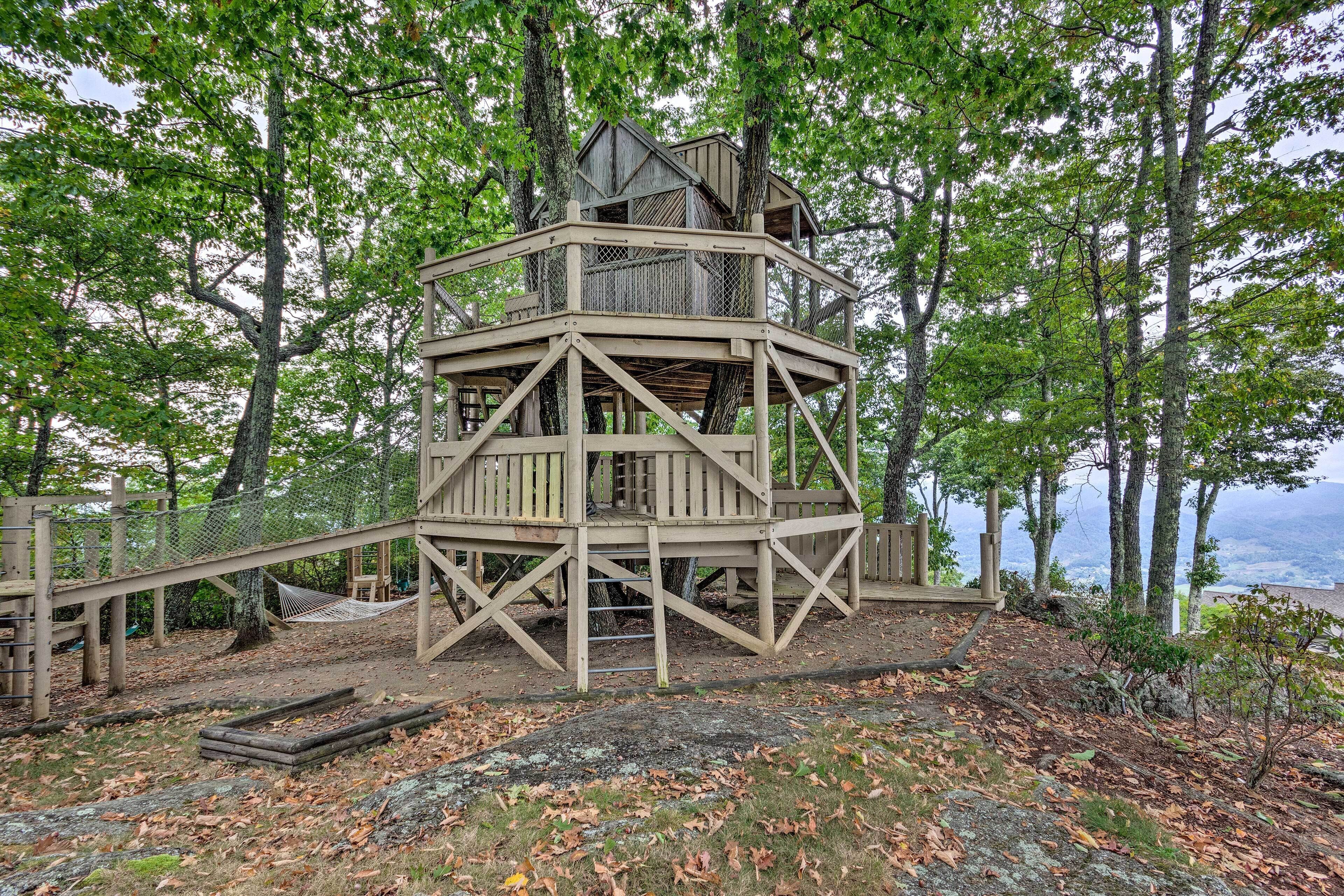 Ein hölzerner Aussichtsturm mit Plattform und Seiltreppe, umgeben von Bäumen und Blick auf die Landschaft.