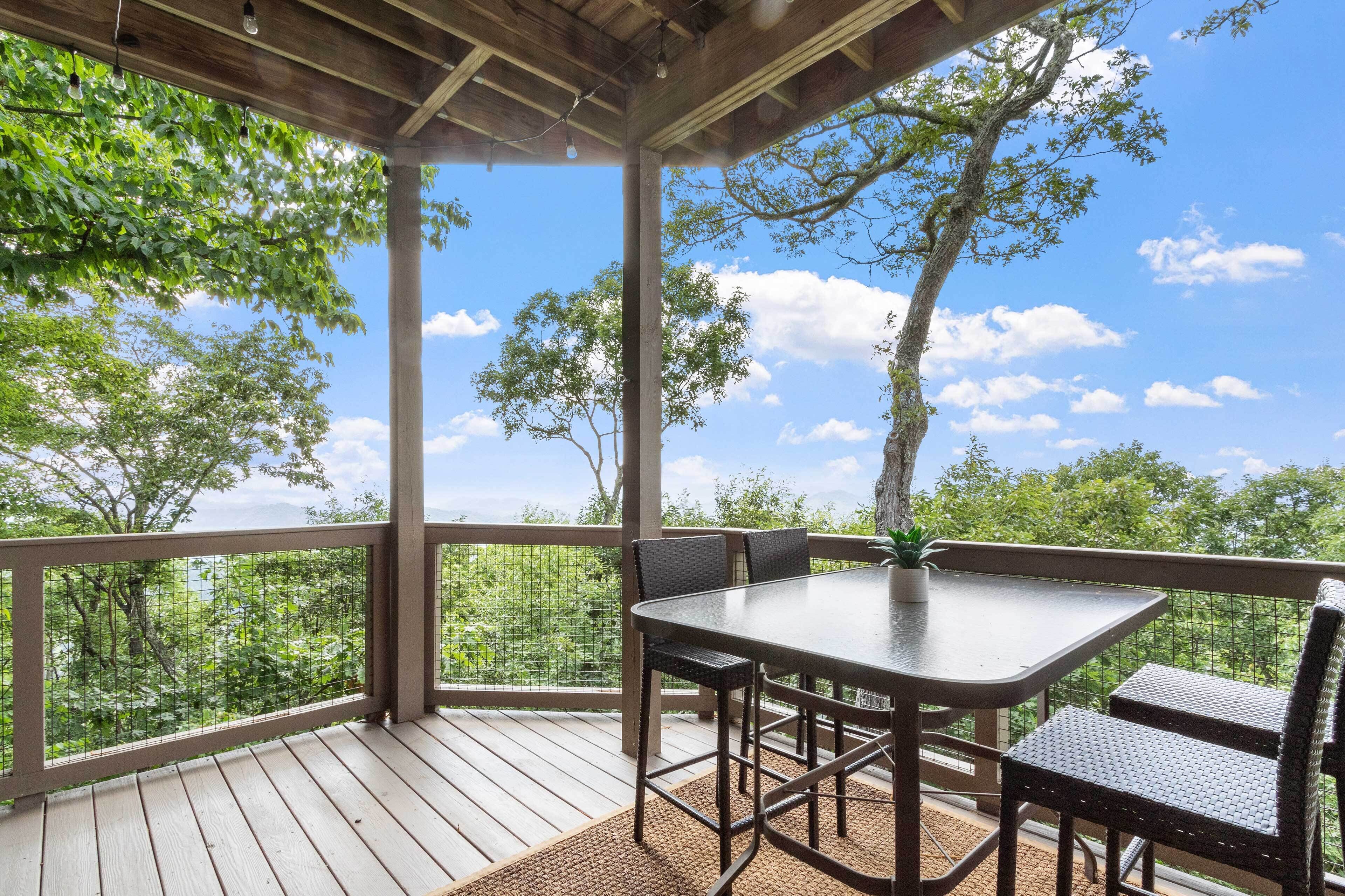 A covered outdoor area with a table and chairs, surrounded by a wooden deck and a metal railing, overlooking a forested area.