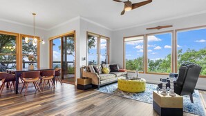 A spacious living area with a wooden floor, a sofa, a dining table with chairs, and large windows offering a view of the outdoors. - Hiking On-site: Mtn-view Burnsville Condo! (Burnsville)