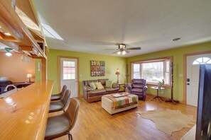 A spacious living room with a wooden dining table, leather chairs, a sofa with cushions, a ceiling fan, and large windows.