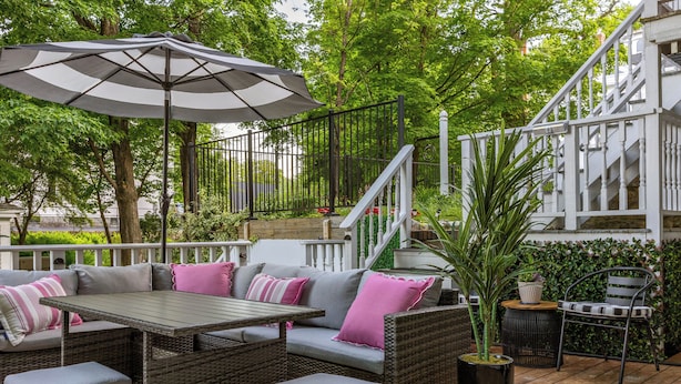 Ein Patio-Bereich mit Korbmöbeln, einem Tisch und einem Sonnenschirm. Im Hintergrund ist eine Treppe mit weißen Geländern und ein grüner Heckenstreifen zu sehen.