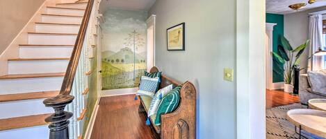 Una escalera de madera con barandilla, un banco con cojines, un mural en la pared y vistas a una sala de estar con una planta y un sofá.