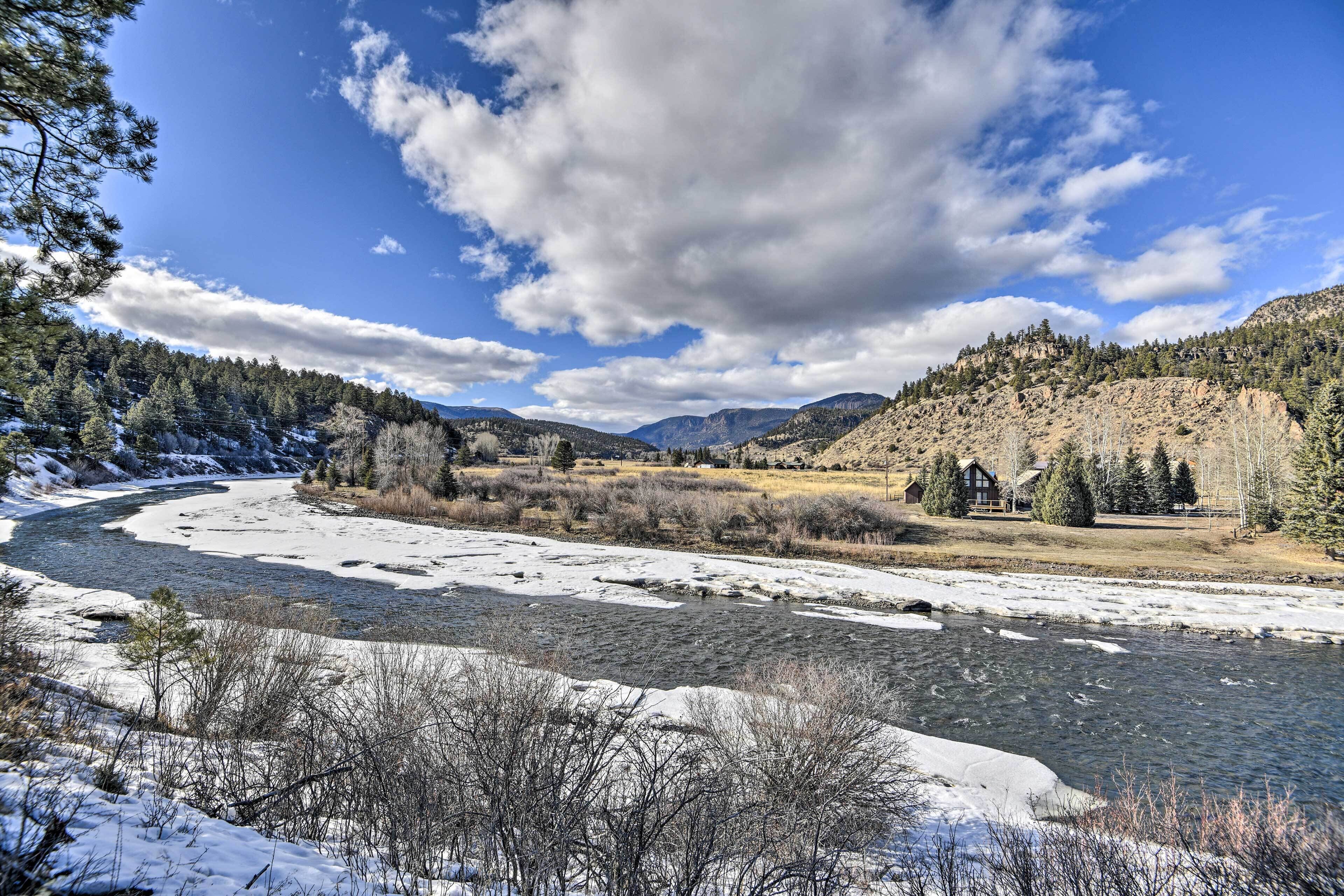 A river with snowbanks, a house with a wooden deck, and a forested hillside.