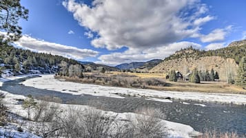 A river with snowbanks, a house with a wooden deck, and a forested hillside.