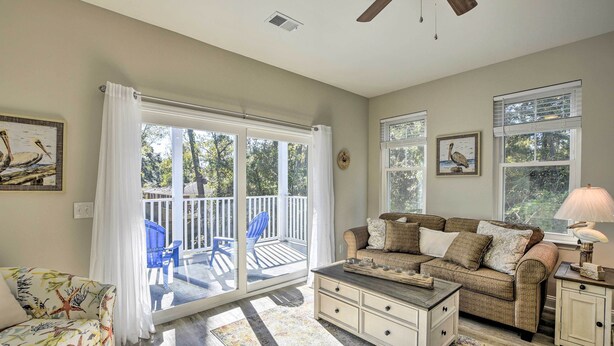 A living room with a sofa, a coffee table, and a lamp. There is a sliding glass door leading to a balcony with chairs. - Modern Escape in the Heart of Murrells Inlet (Murrells Inlet)