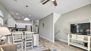 A modern kitchen with white cabinets, a dining area with a white table and chairs, a TV on a white stand, and a ceiling fan. - Modern Escape in the Heart of Murrells Inlet (Murrells Inlet)