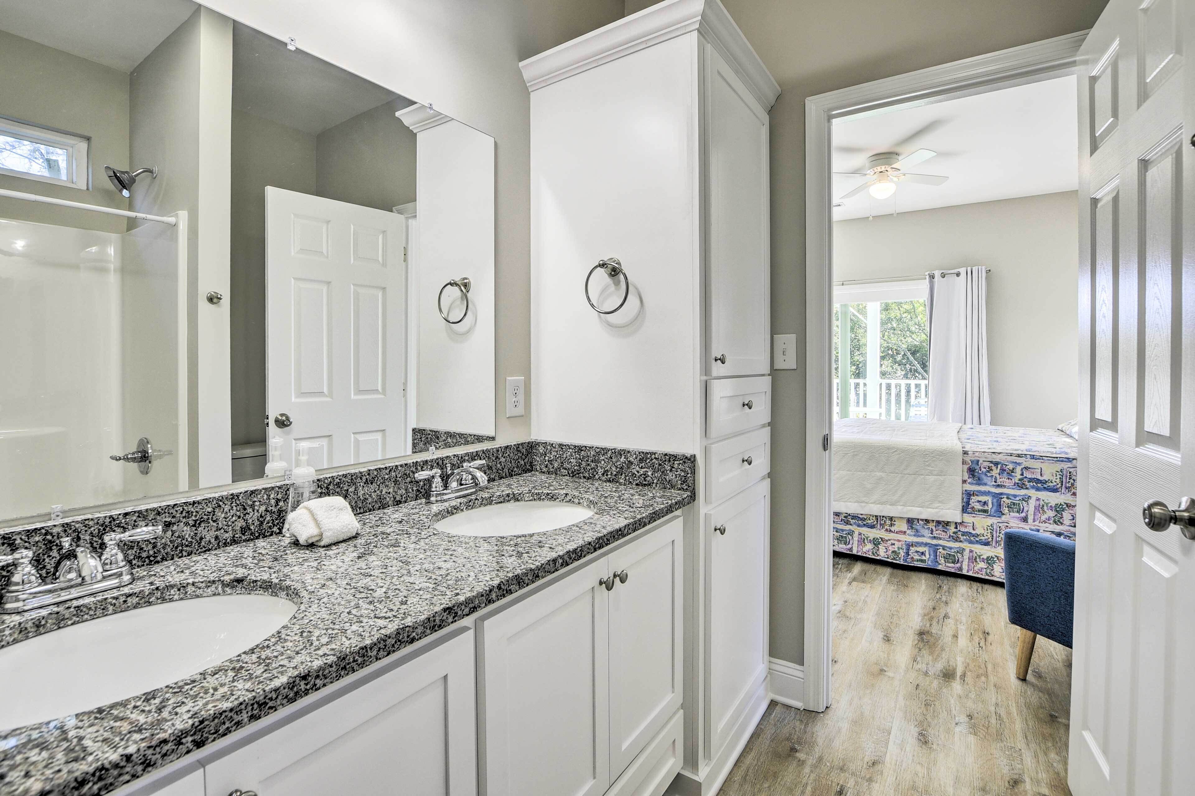A bathroom with two sinks, a large mirror, and a door leading to a bedroom with a bed and a view of the outdoors.