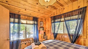 A wooden cabin bedroom with a plaid bedspread, two windows with curtains, a bedside lamp, and a framed picture on the wall. - California Retreat w/ Deck, Fire Pit & Grill! (Long Barn)
