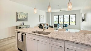 A modern kitchen with a granite countertop, stainless steel sink, and white cabinetry. There are pendant lights, a dining area with a table and chairs, and a large window offering a view of the outside. - Lakeside Montgomery Condo Near Boat Rental! (Montgomery)