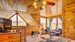 Ein gemütliches Blockhaus-Interieur mit Holzbalkendecke, einem Bett mit gesteckter Bettdecke, einem Schreibtisch mit Fernseher, einem Ledersessel und Blick ins Freie.