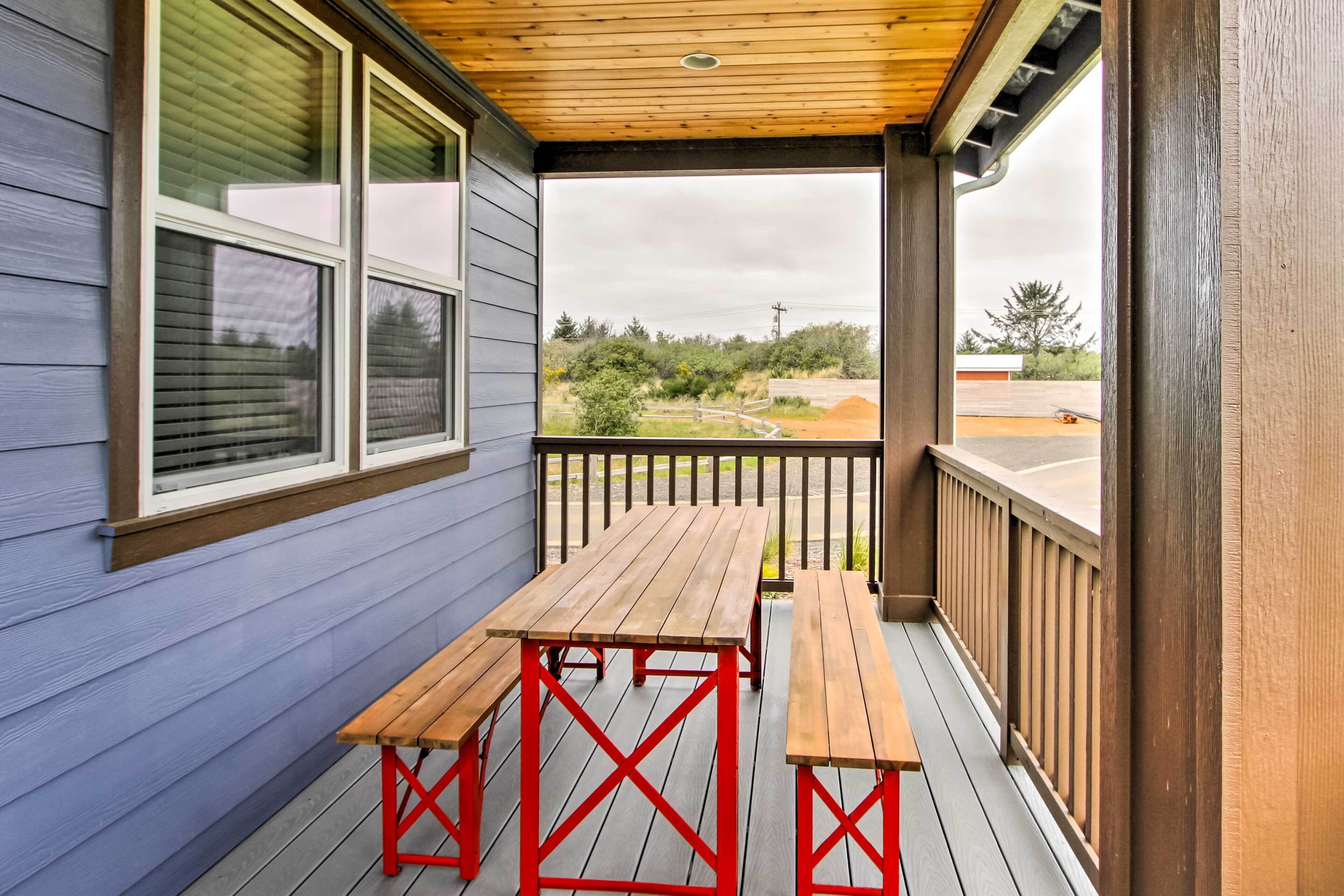 A covered porch with a wooden table and benches, a view of the surrounding landscape, and a building with blue siding.