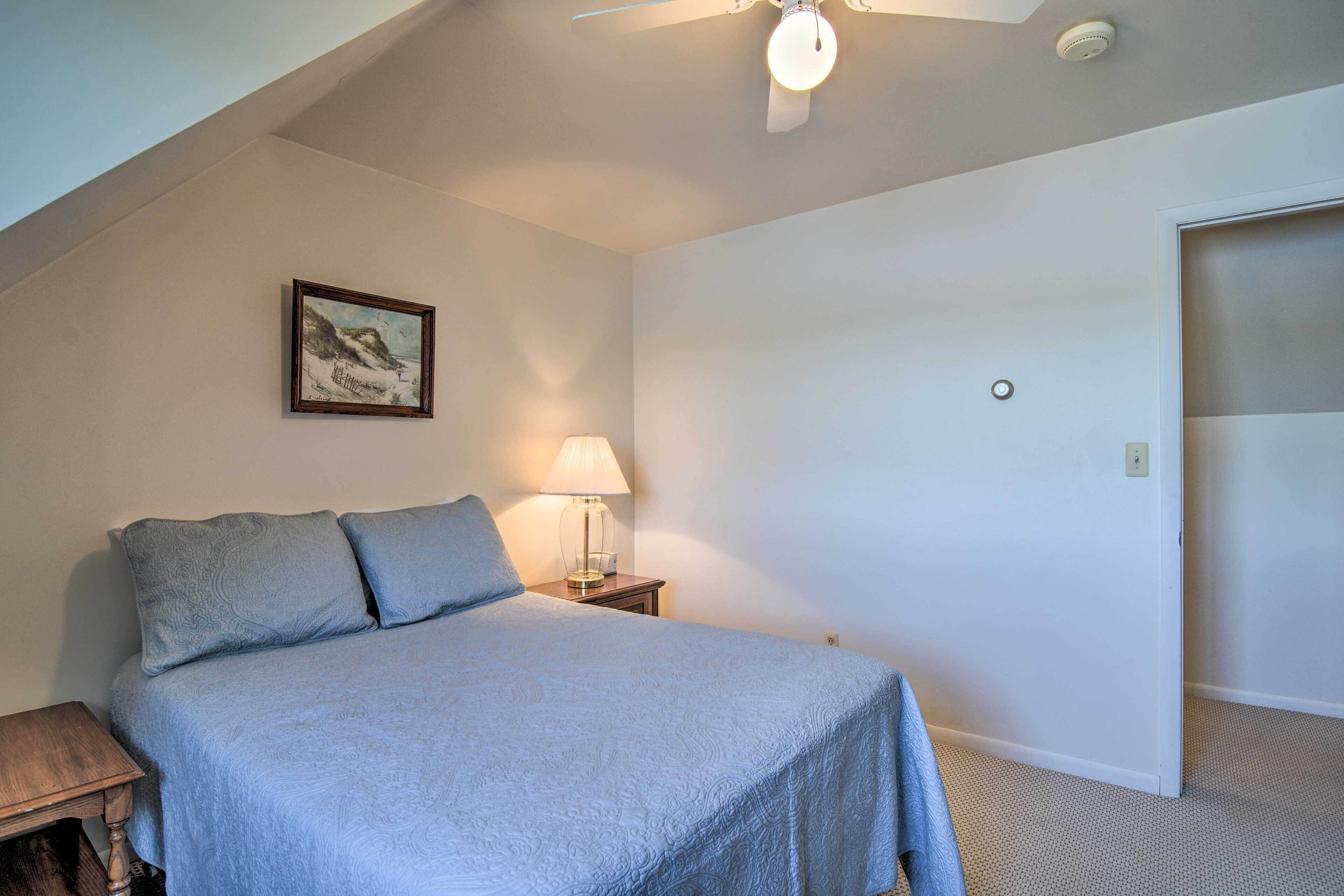 A bedroom with a bed, a nightstand, a lamp, a ceiling fan, a painting, and a door.