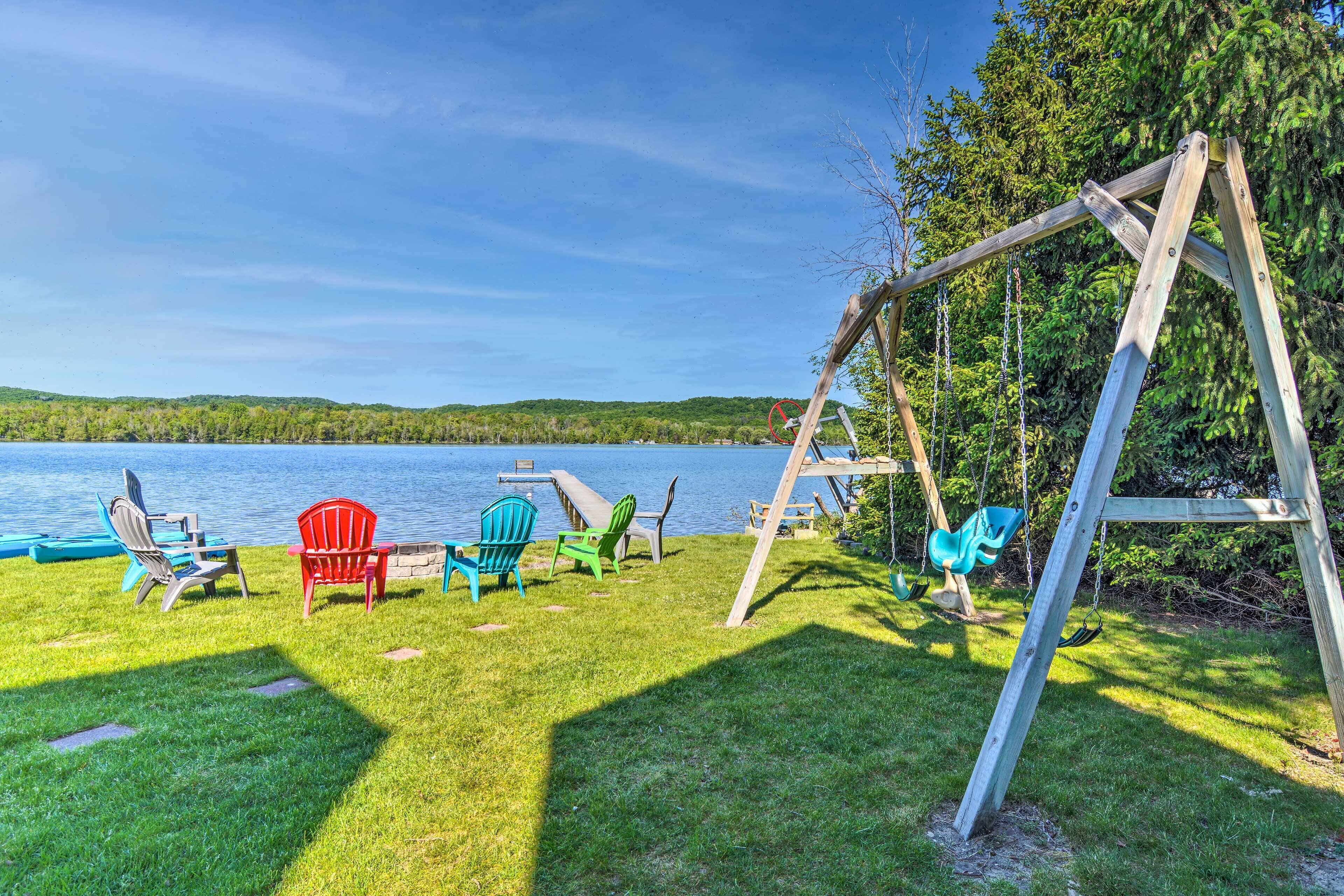 A wooden swing set and Adirondack chairs by a lake.